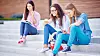 Modern teen girls with cellphones sitting on stairs on background of reading student