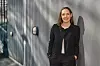 smilende kvinne lener seg nonchalant mot en murvegg med designerutelamper. Det må være en vakker dag, for sollys som sildrer inn mellom lameller i bakgrunnen får henne til å kaste skygge på den hvite murpussen.
