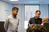 To menn står i et kontor mens flere personer deltar i et møte.