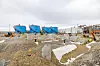 Industriområde under bygging med blå tanker, grusdekke, betongfundamenter og anleggsmaskiner under en overskyet himmel.