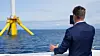 Energiminister Terje Aasland, sett bakfra i i mørk dress, fotograferer en gul flytende vindturbin.