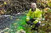 En person i gul arbeidsjakke kneler ved en bekk med grønt fargestoff i vannet.