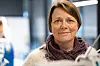 Kvinne med kort brunt hår og skjerf ser mot kamera i et lyst innendørs arbeidsmiljø.