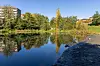 Åpent vannløp med speilblankt vann, trær, gress og boligbygg i bakgrunnen.