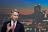 Mann i mørk dress og rødt slips taler foran en belyst bysilhuett ved vannet.